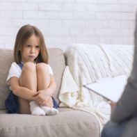 Child Counseling Concept. Concerned Little Girl At Therapy Session With Children Psychologist Sitting With Her Knees Up On Couch