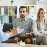 Offended woman with crossed arms looking aside while her husband trying to explaining their problem to counselor